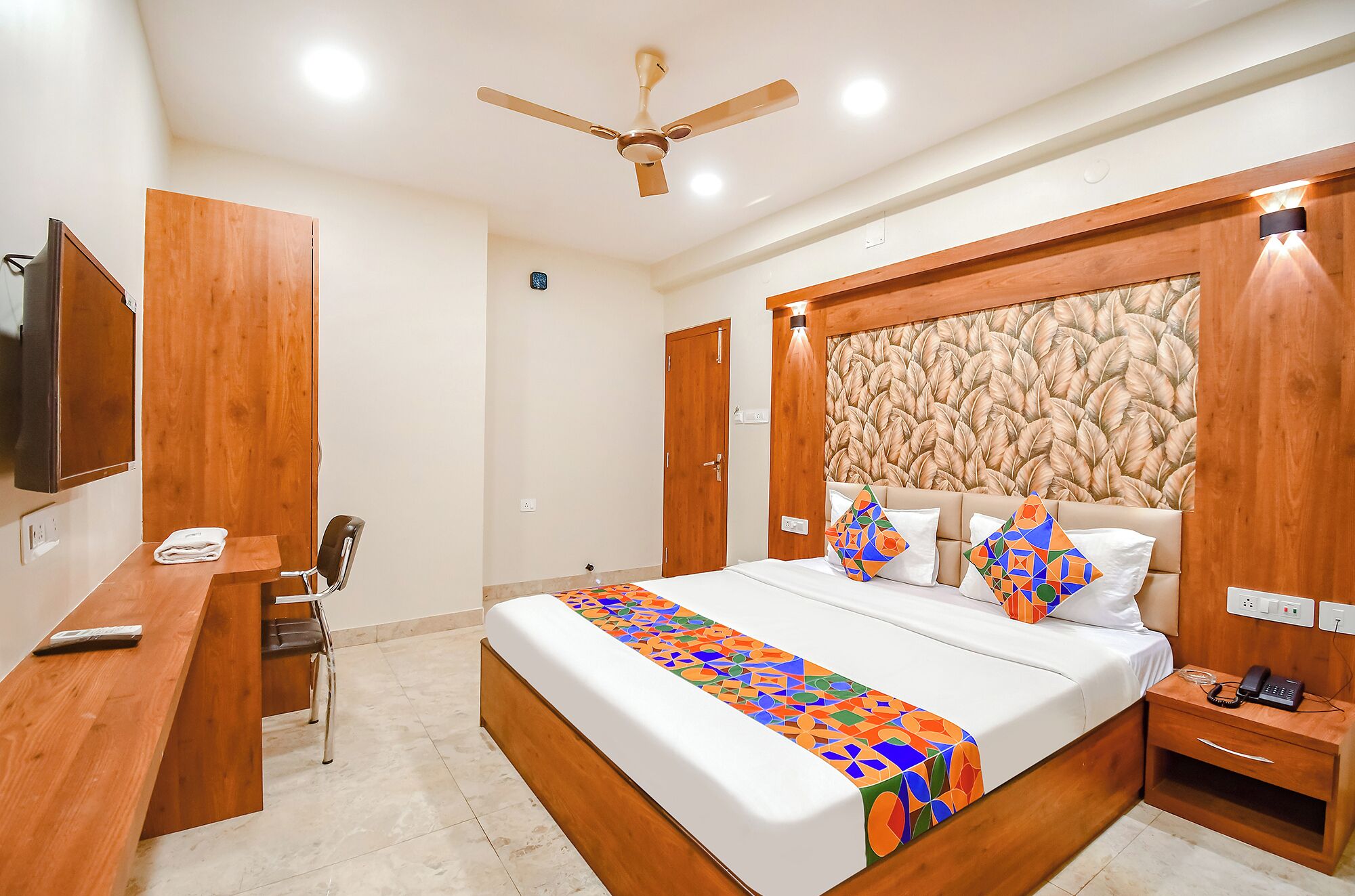 A modern bedroom with a wooden headboard, a bed with colorful bedding, a desk with a chair, and a ceiling fan.