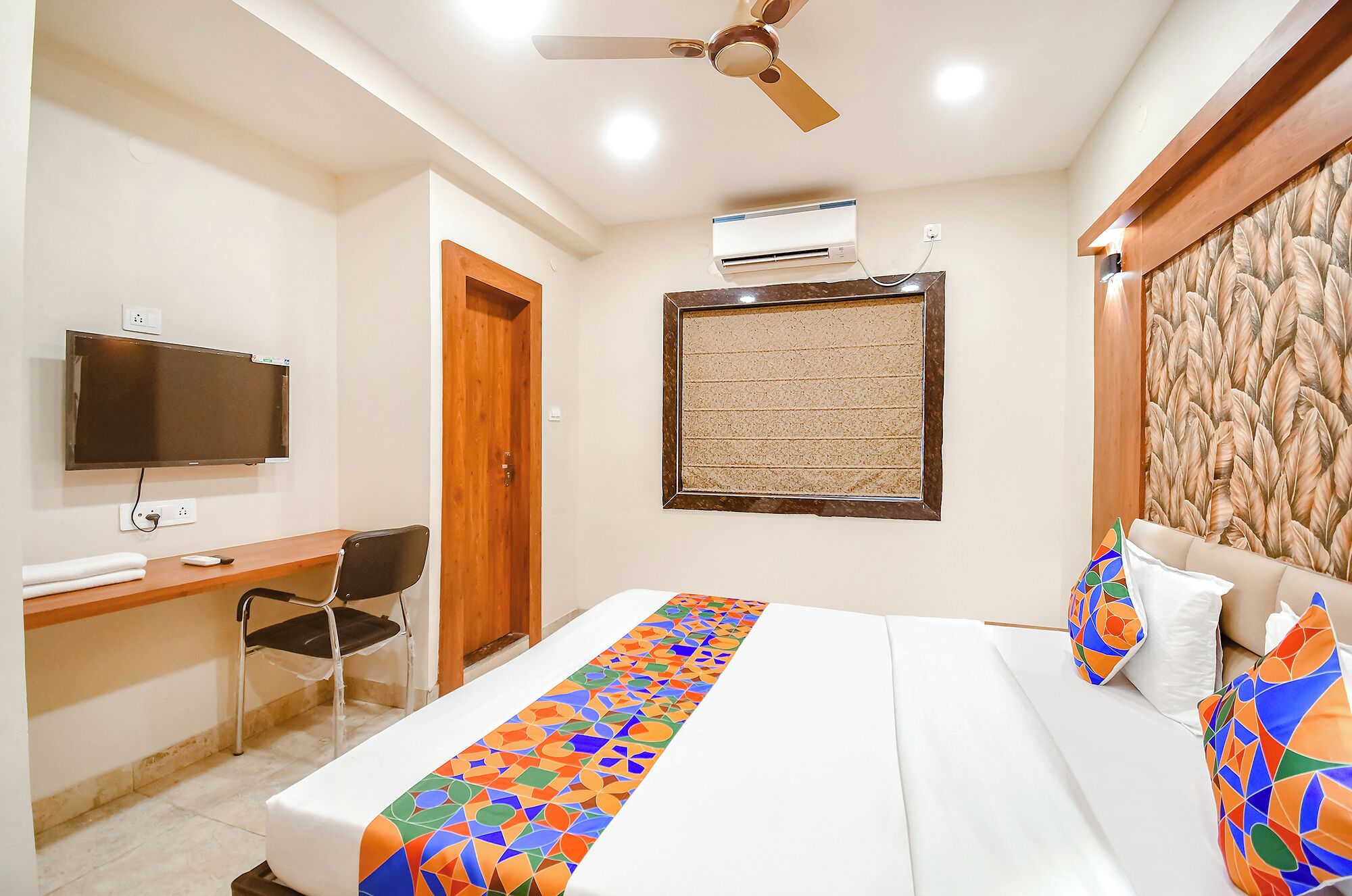 A hotel room with a bed, a desk, a chair, a television, an air conditioning unit, and a ceiling fan.