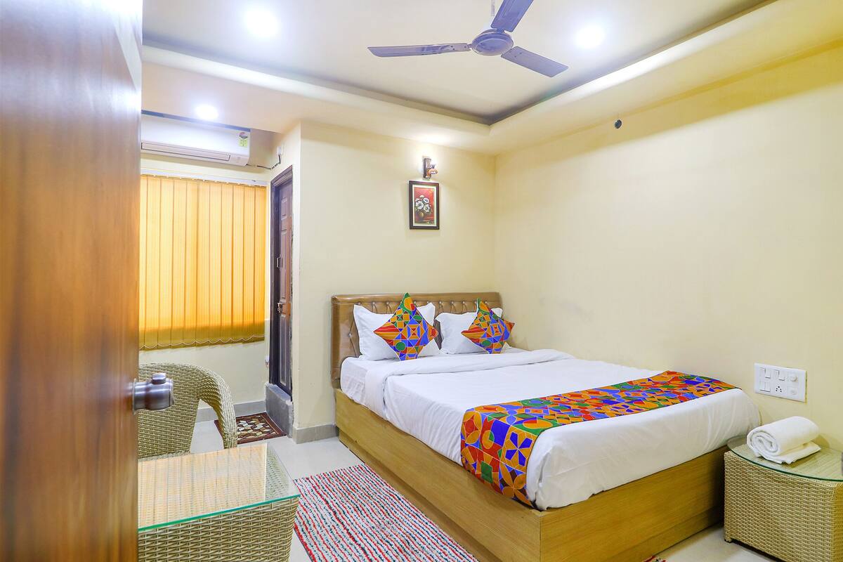 A hotel room with a bed, a ceiling fan, a wall-mounted air conditioner, a small table, and a wicker basket.