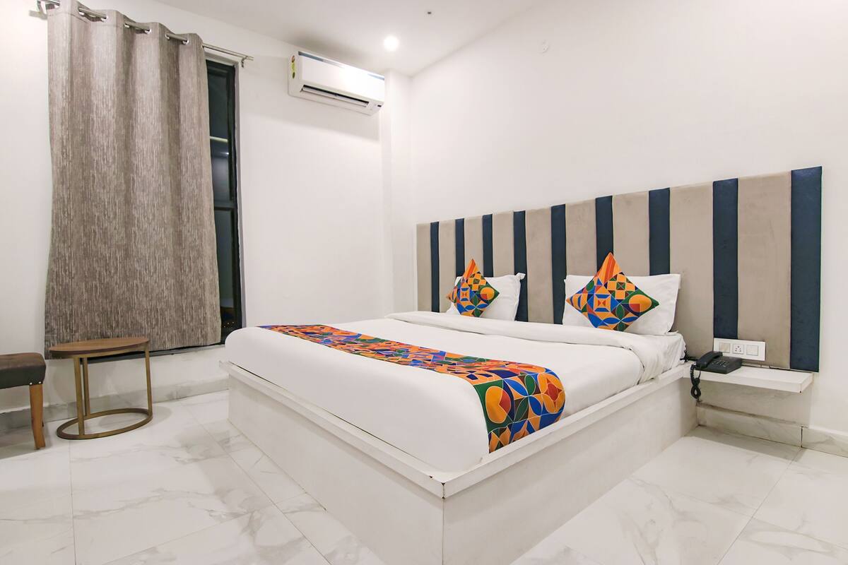 A hotel room with a bed, a bedside table with a phone, a small table, a curtain, and an air conditioning unit.