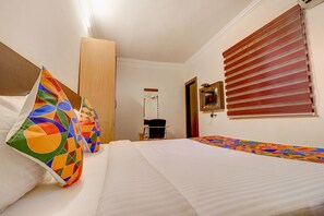 Quarto de hotel com cama, almofadas coloridas, uma cadeira, escrivaninha e janela com persianas.