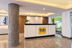 A hotel reception area with a modern design, featuring a reception desk with a logo, a large pillar, and a view of the outdoors through a glass door.