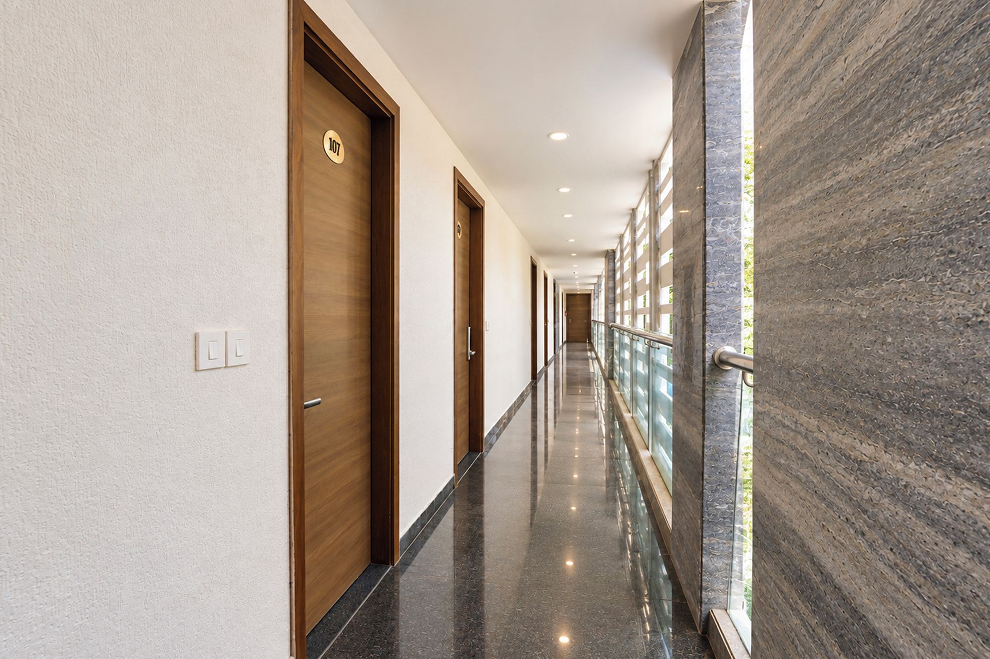Un pasillo con puertas de madera, un muro de mármol y una barandilla de cristal.