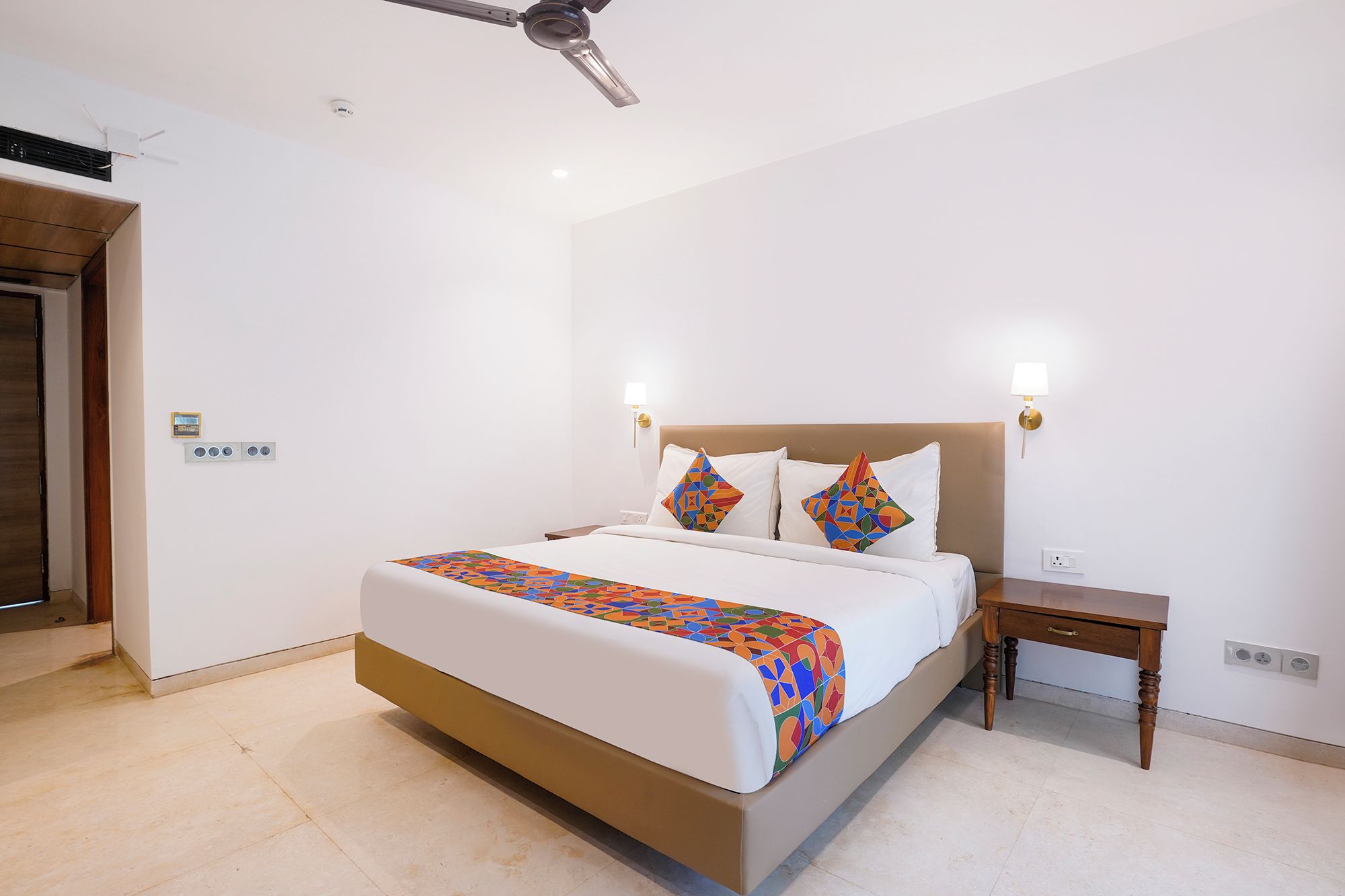 A hotel room with a bed, bedside table, nightstand, wall-mounted lamp, and a ceiling fan.