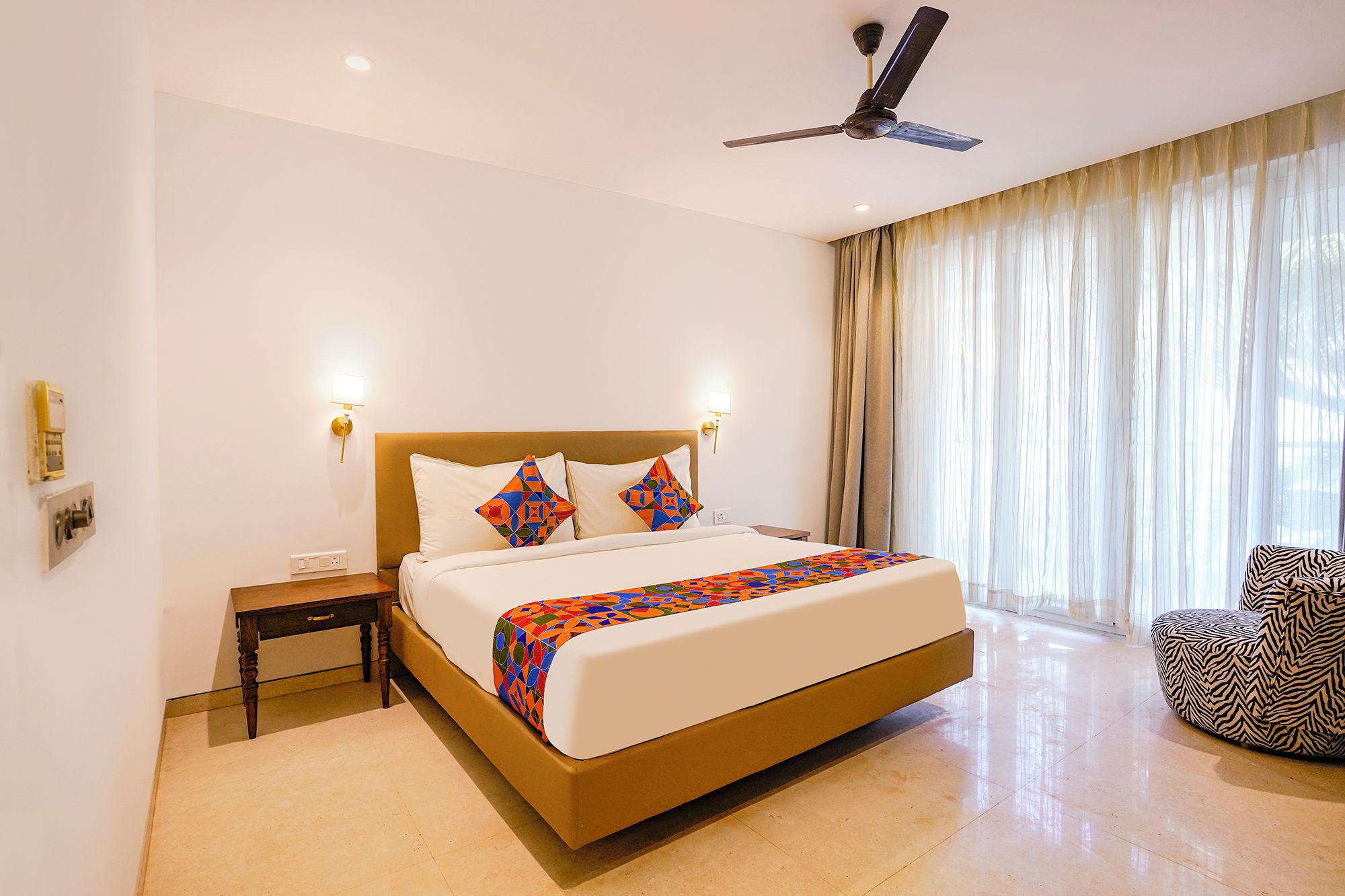 A hotel room with a large bed, a ceiling fan, a nightstand, a chair, and a window with curtains.