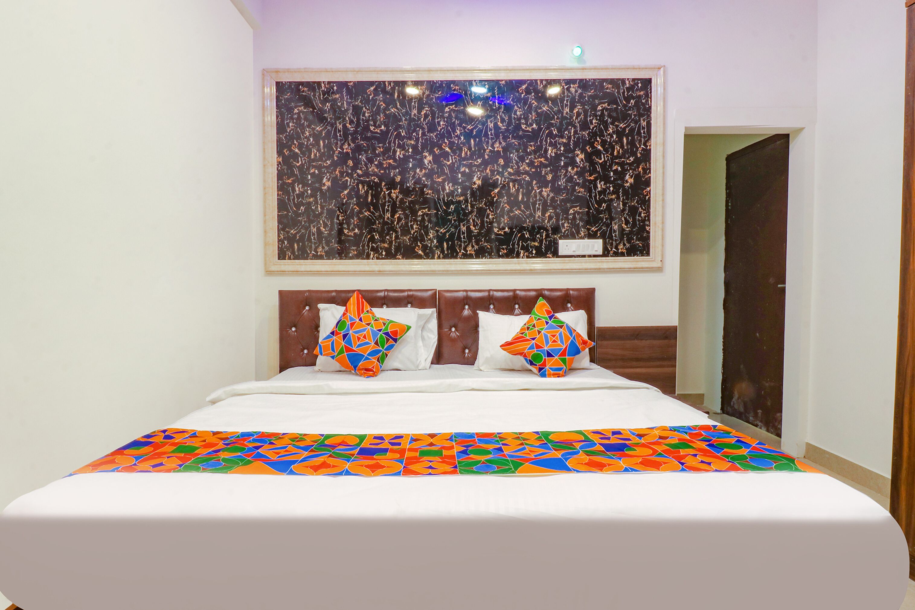 A hotel room with a large bed, a colorful geometric-patterned bedspread, two pillows with geometric patterns, a large abstract painting on the wall, and a wooden headboard.
