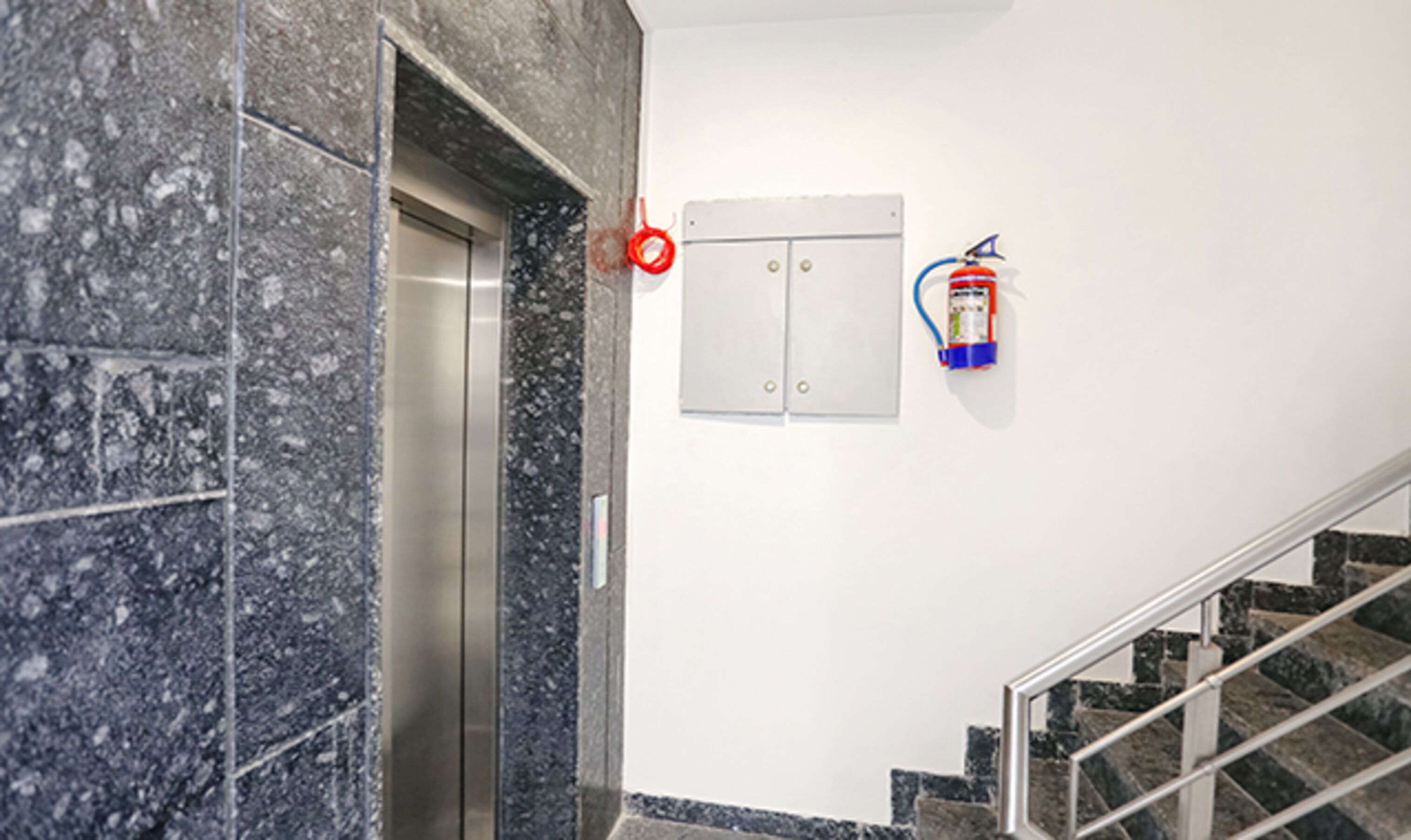 Een lift met een marmeren wand, een brandblusser en een trap met metalen leuningen.