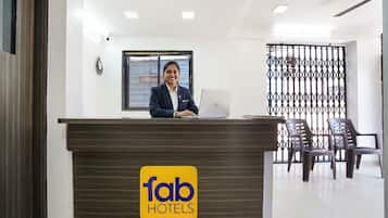A hotel reception desk with a logo, a receptionist, a laptop, and a seating area with chairs.