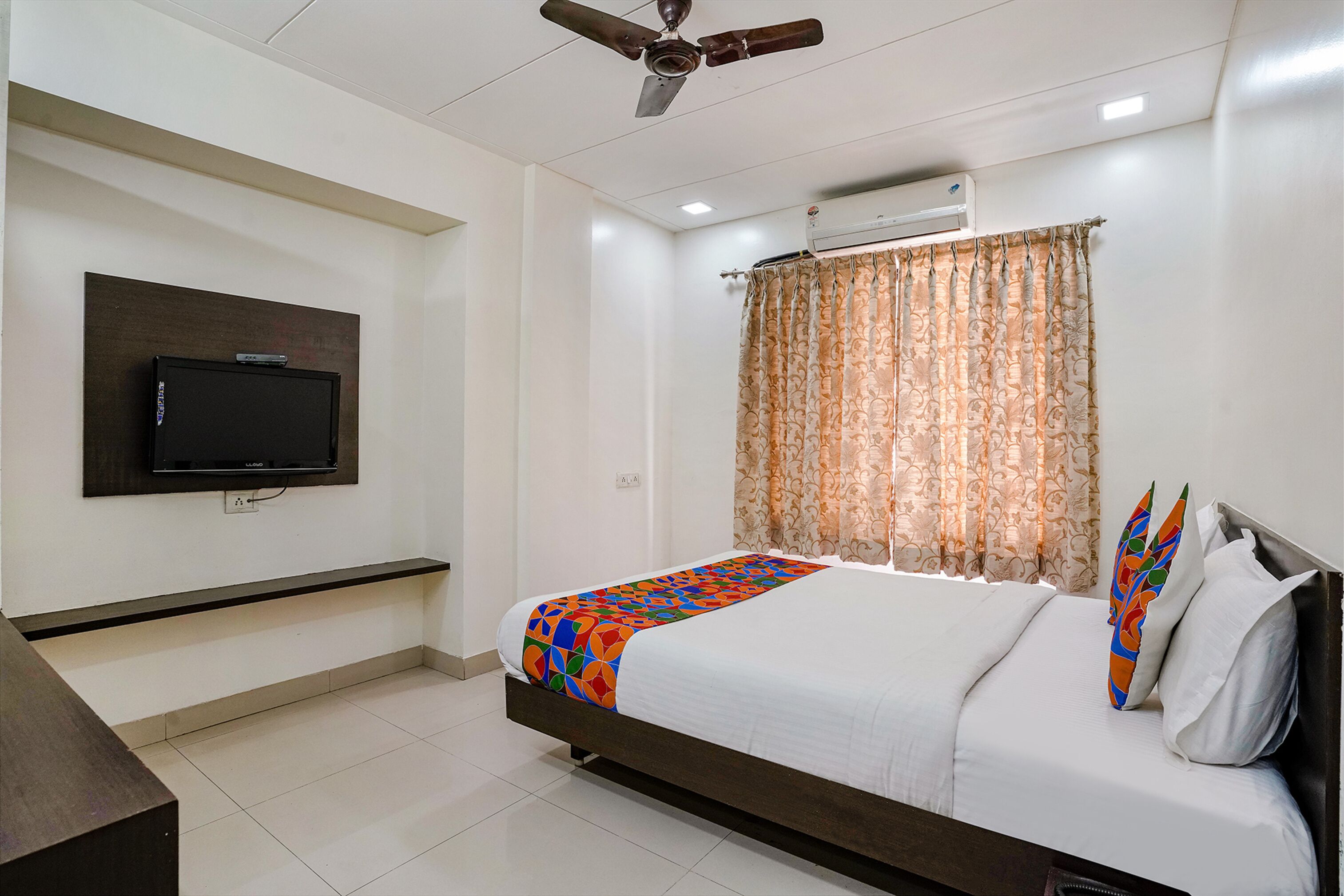 A hotel room with a bed, a TV mounted on the wall, a ceiling fan, and a window with curtains.