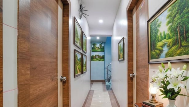 A hallway with wooden doors, framed landscape paintings, a staircase, and a table with books and a vase of flowers. - FabHotel White Gold (Navi Mumbai)