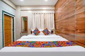 Une chambre à coucher moderne avec une tête de lit en bois, un grand lit recouvert d’une courtepointe aux motifs géométriques colorés, deux coussins décoratifs et un miroir au-dessus du lit.