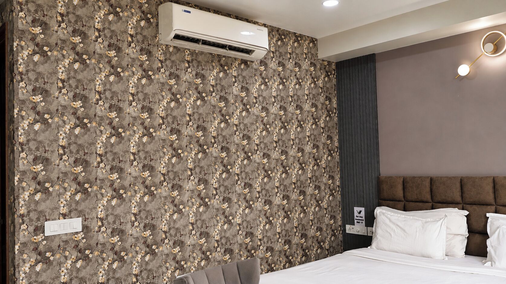 A hotel room with a floral wallpaper, a bed with white linens, a wall-mounted air conditioner, and a modern light fixture.