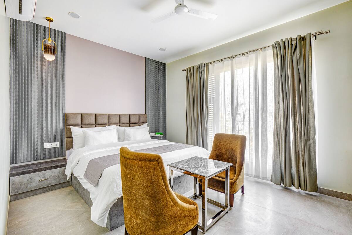 Een hotelkamer met een bed, twee stoelen, een klein tafeltje, een plafondventilator en een raam met gordijnen.
