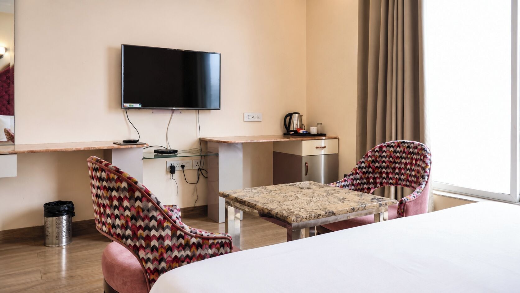A hotel room with a bed, a desk, a television, and two patterned chairs.