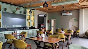 L’intérieur d’un restaurant, avec des tables et des chaises en bois, un espace cuisine et des plantes décoratives.