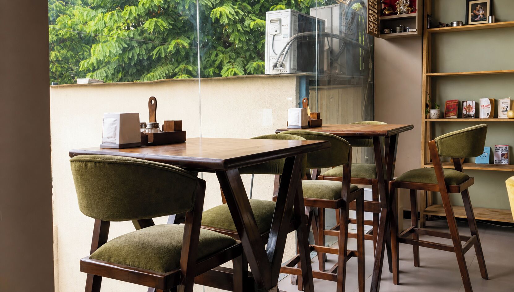 A dining area with a table and chairs, a window with a view of greenery, and a bookshelf with various items.
