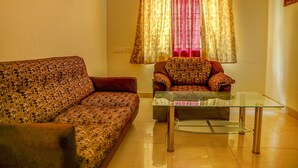 A living room with a sofa and armchair, a glass coffee table, and patterned curtains. - Fabhotel Hibiscus Stays (Chennai)