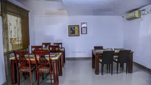 A dining area with wooden tables and chairs, a window with a curtain, and an air conditioning unit on the wall. - Fabhotel Hibiscus Stays (Chennai)