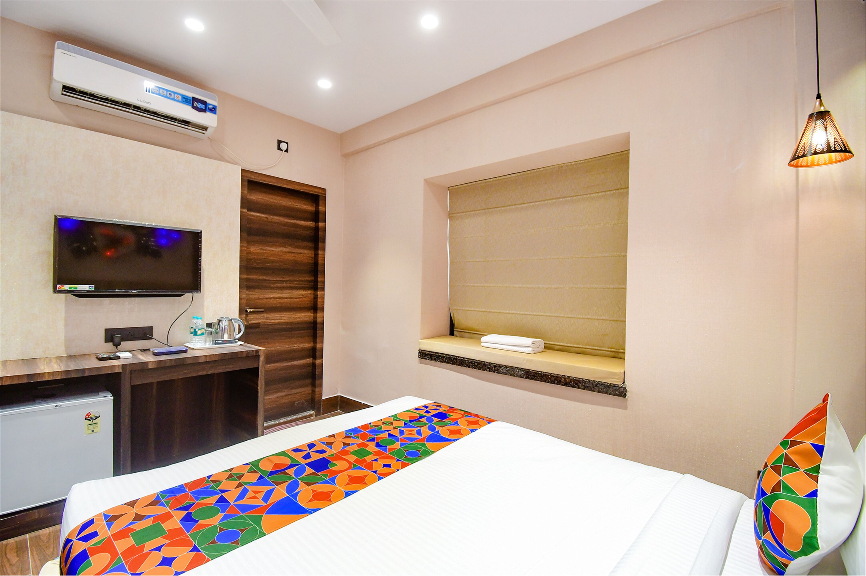 A hotel room with a bed, a TV, a desk with a kettle, and an air conditioner.