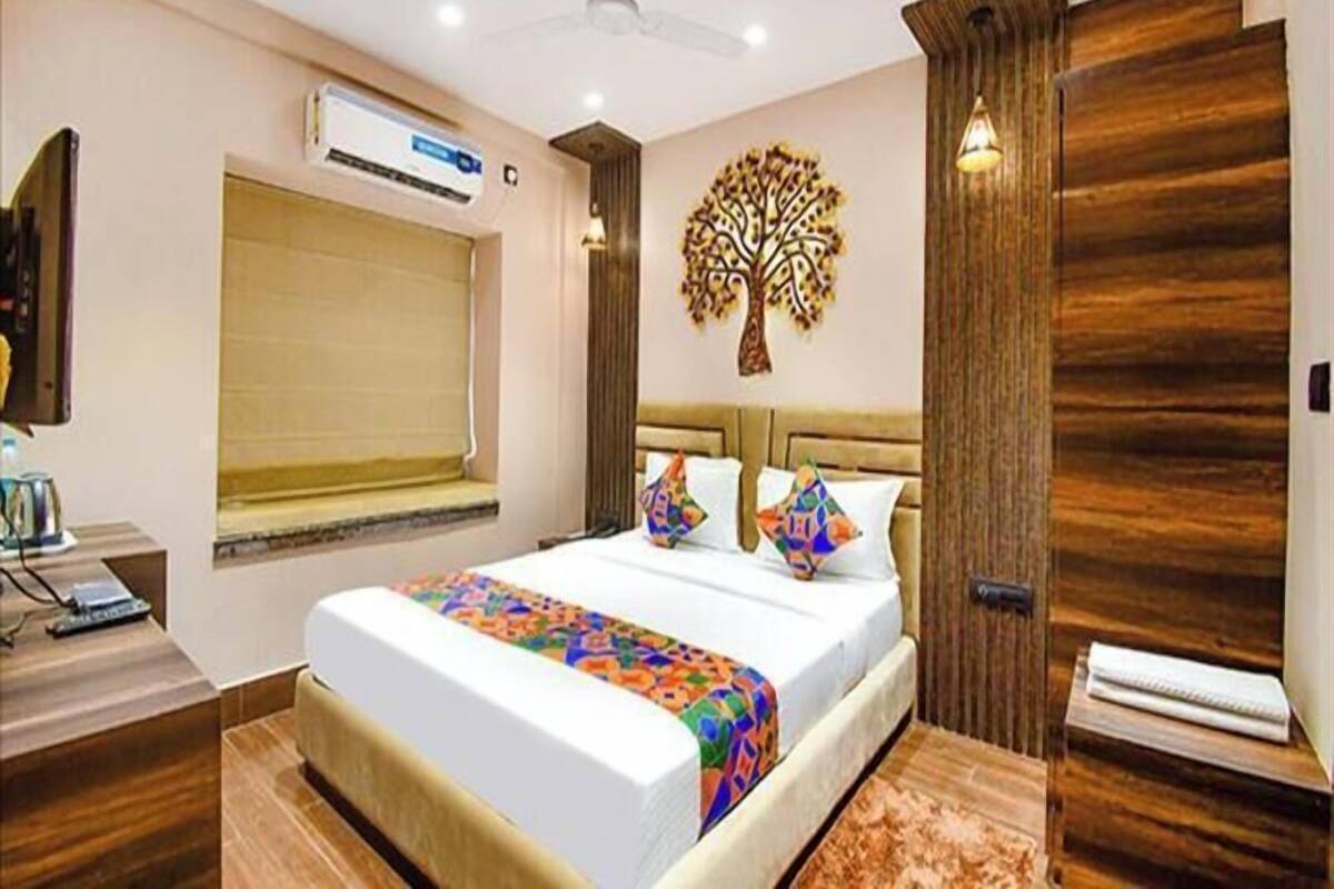 A hotel room with a bed, a window with a blind, a wall-mounted air conditioner, a ceiling fan, and a decorative tree wall art piece.
