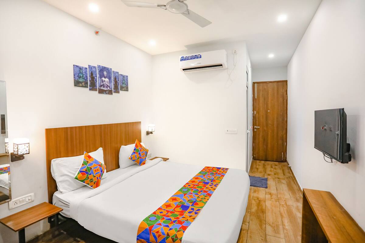 Habitación de hotel con cama, cabecera de madera, ventilador de techo, unidad de aire acondicionado, puerta de madera, televisor y una mesita.