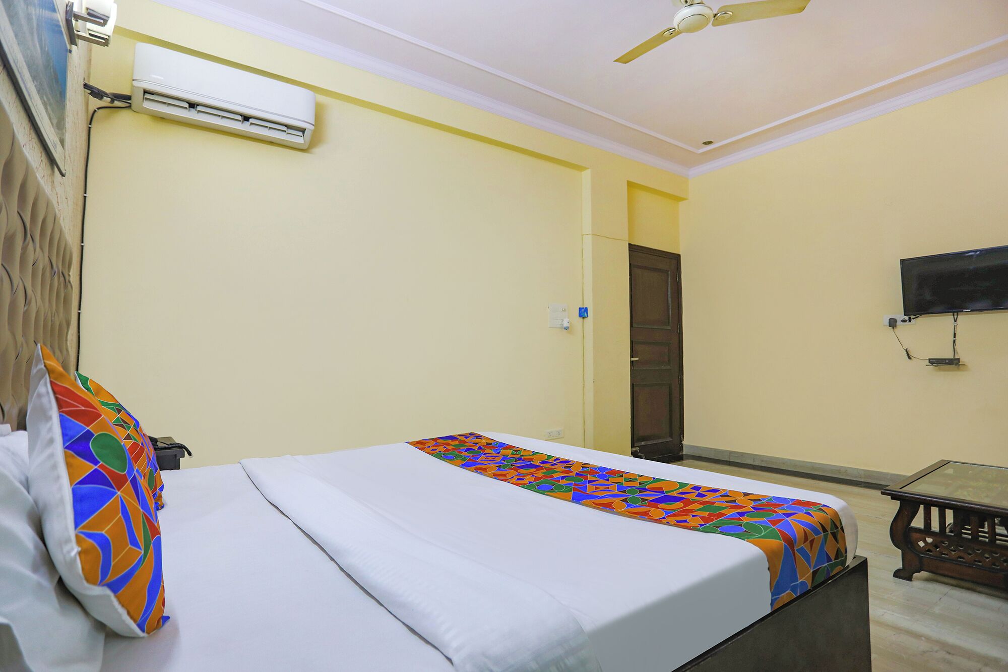 A hotel room with a bed, a ceiling fan, an air conditioning unit, a TV mounted on the wall, and a wooden table.