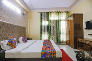 A hotel room with a tufted headboard, a large bed with a colorful geometric bedspread, a wooden wardrobe, and a television on the wall. - Fabexpress Hm Palace (Noida)