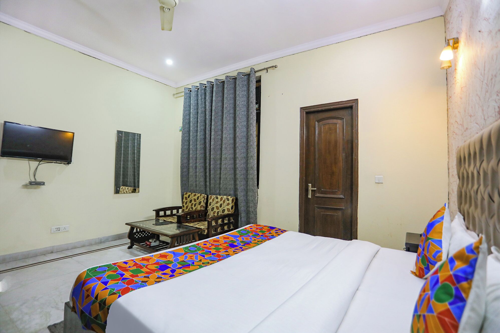 Habitación de hotel con cama, televisión montada en la pared, puerta de madera y ventana con cortinas.