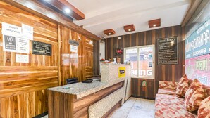 A hotel reception area with wooden paneling, a reception desk, a sofa with patterned cushions, and a digital screen displaying information. - Fabhotel Royal Aanandam (Gwalior)