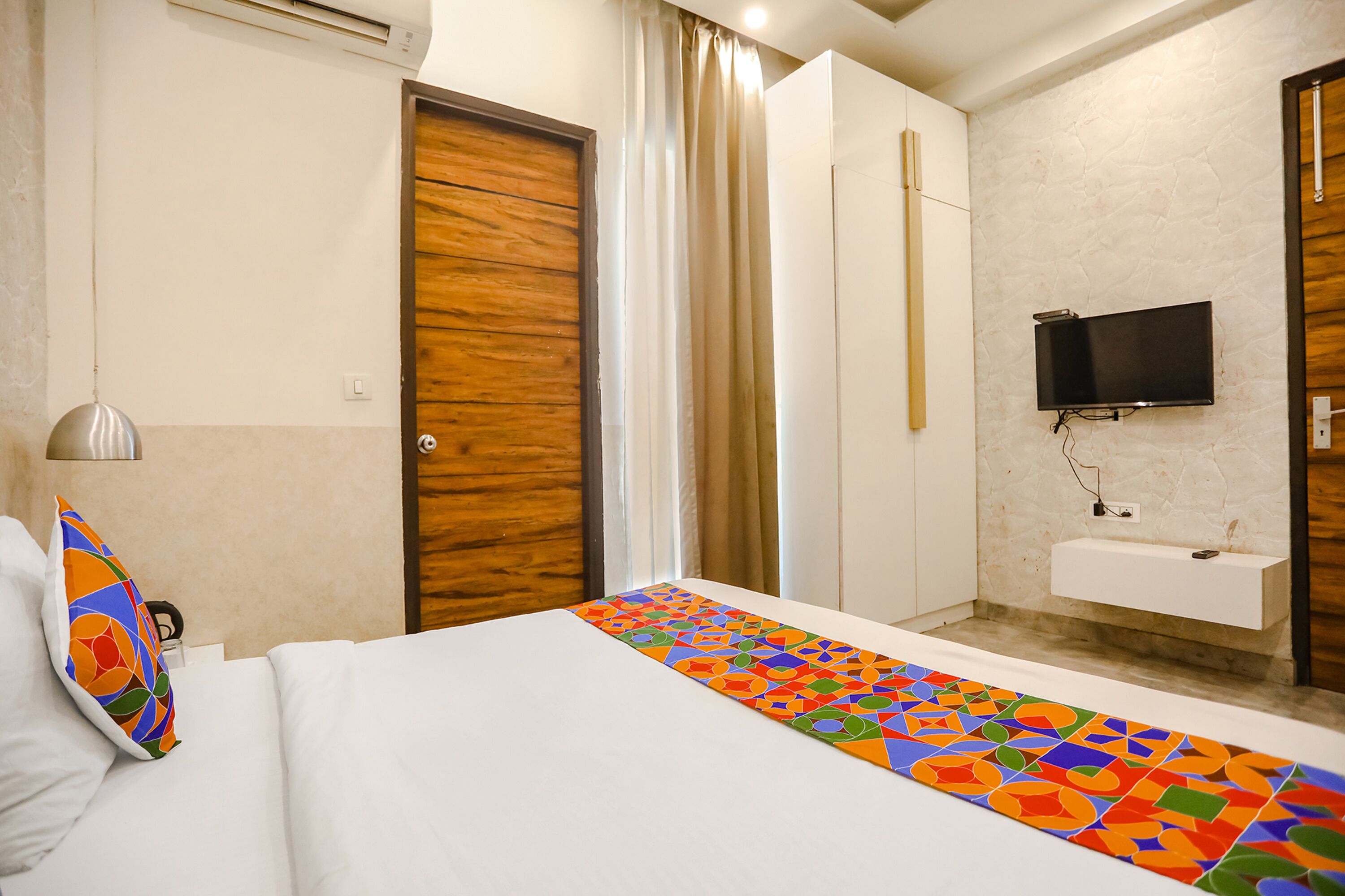 A hotel room with a bed, a TV mounted on the wall, a bathroom with a sink, and a window with curtains.