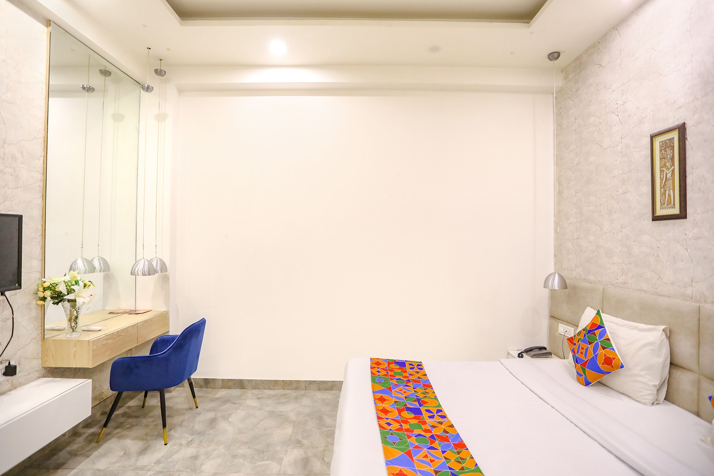 Habitación de hotel con cama, escritorio con silla, espejo y mesita de noche con lámpara.