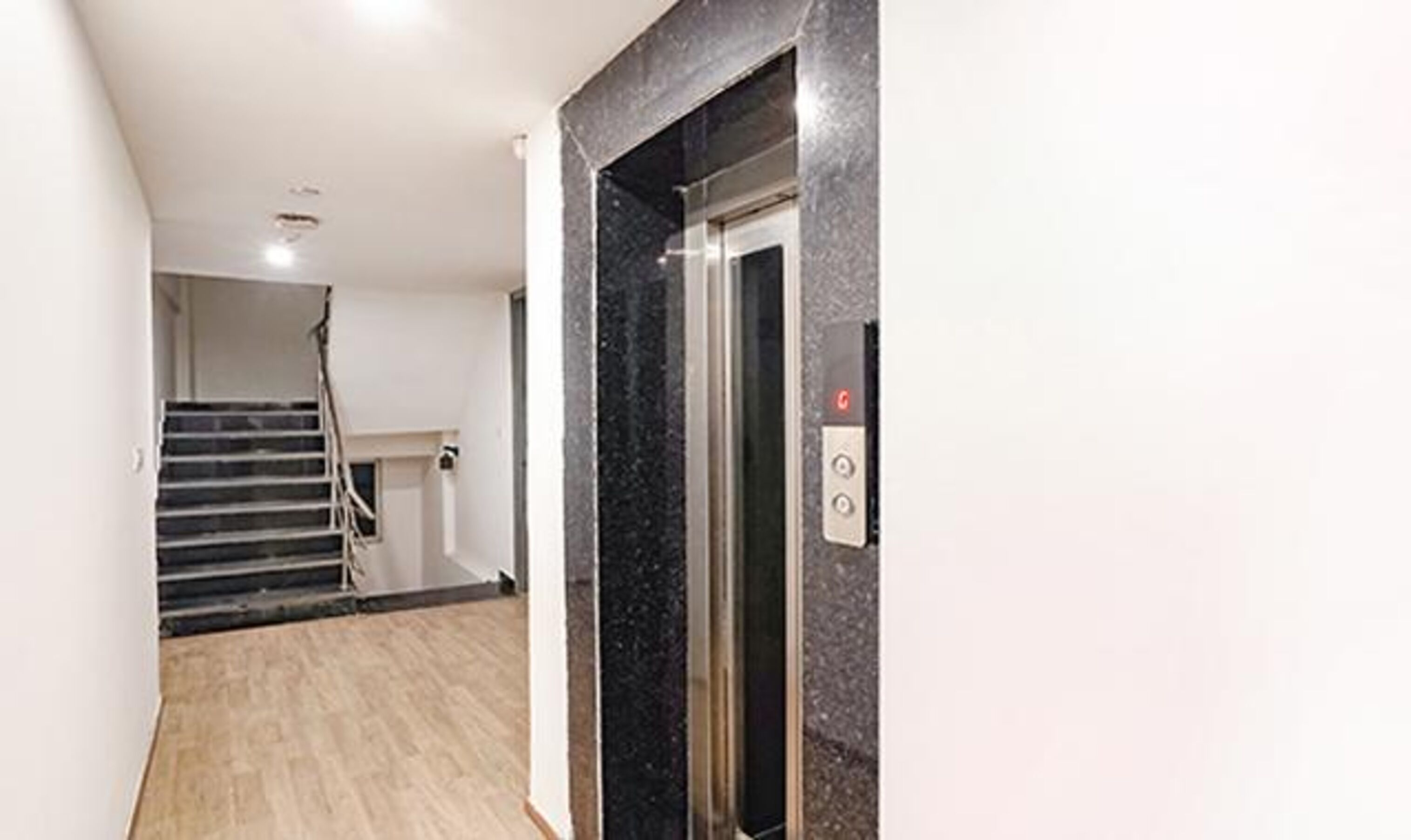 A modern elevator with a marble finish, a staircase with a handrail, and a light-colored wooden floor.