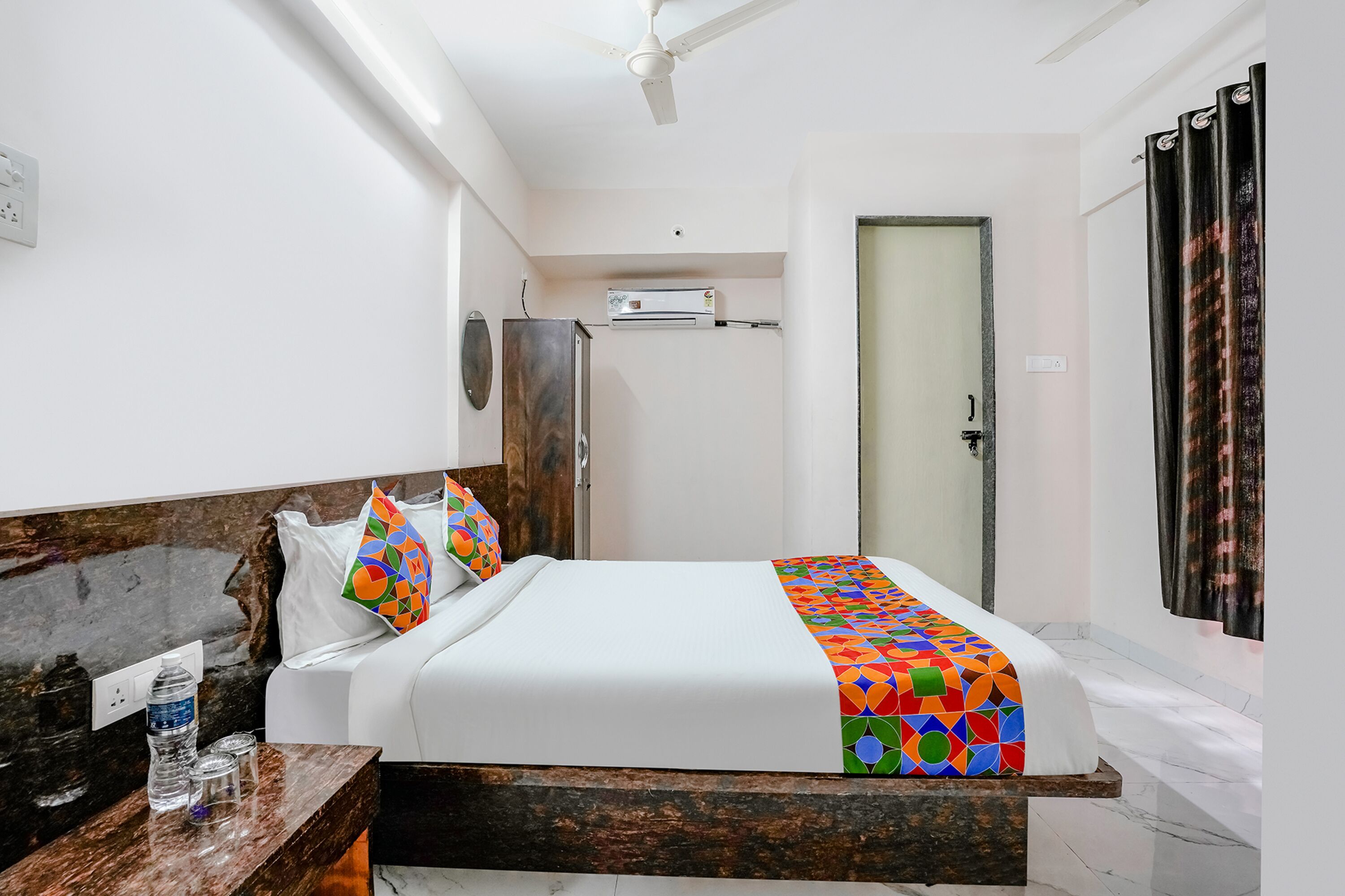 Une chambre d’hôtel avec un lit, une tête de lit en bois, un sol en marbre, un ventilateur de plafond et une porte avec un rideau.