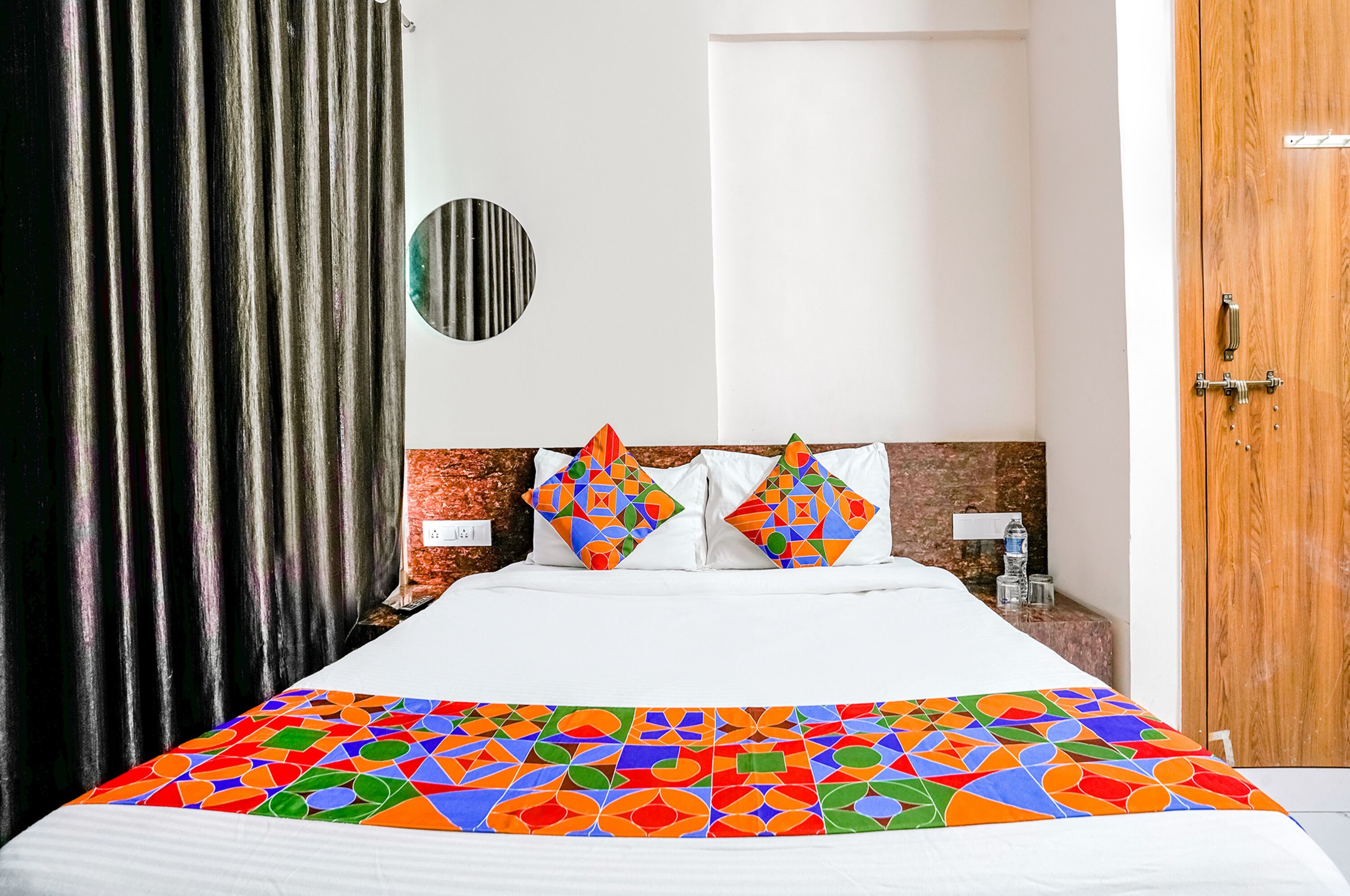 A hotel room with a bed, a wooden headboard, a round mirror, and a door.