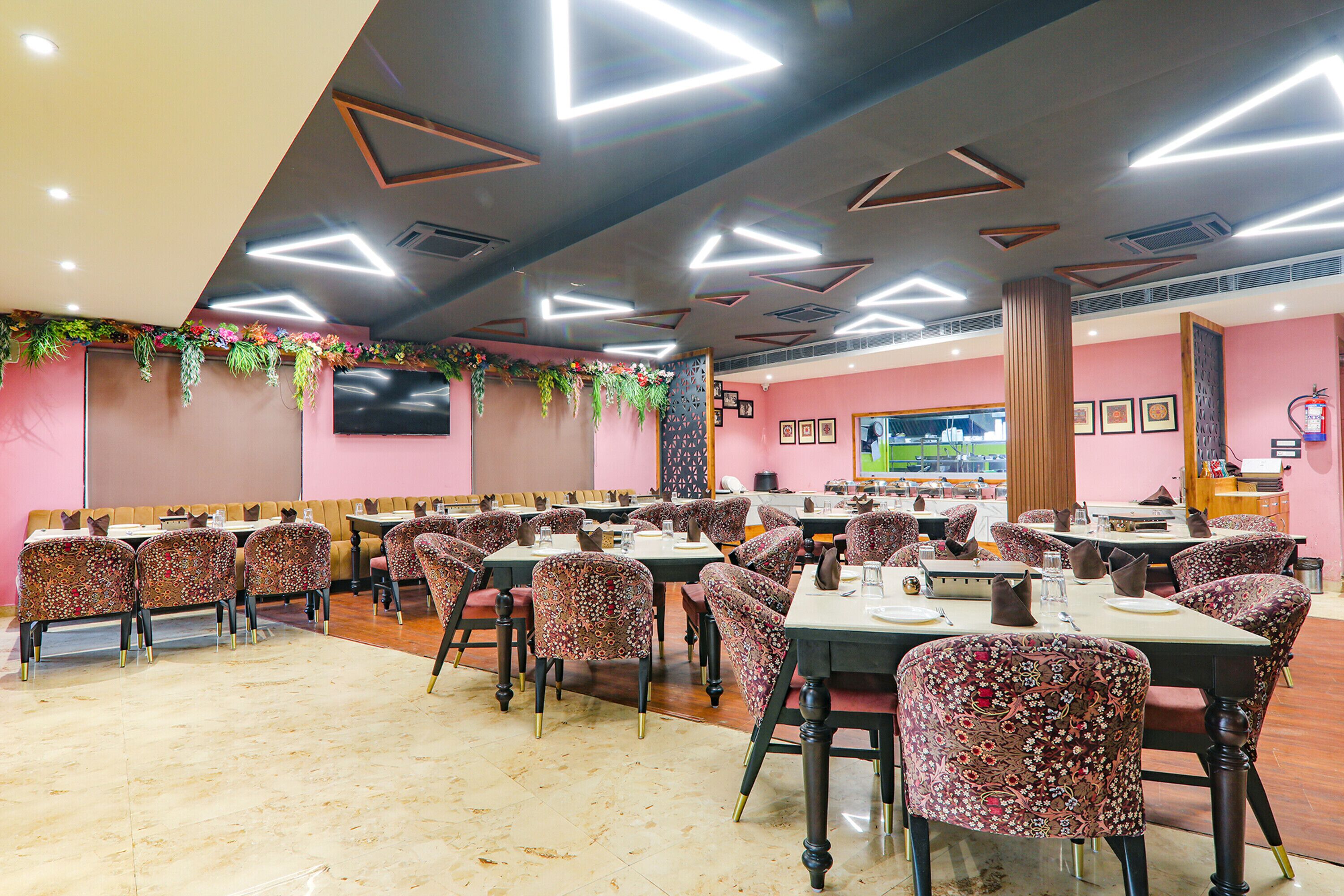 A restaurant with multiple tables and chairs, a flat-screen TV mounted on the wall, and decorative plants on the ceiling.