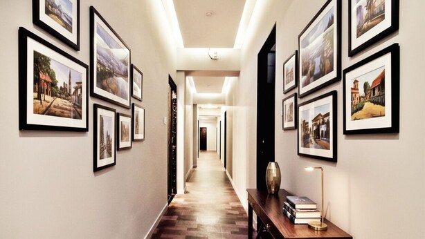 A hallway with framed pictures on the walls, a wooden floor, and a desk with a lamp and books. - FabHotel Pratiksha (Ahmedabad)
