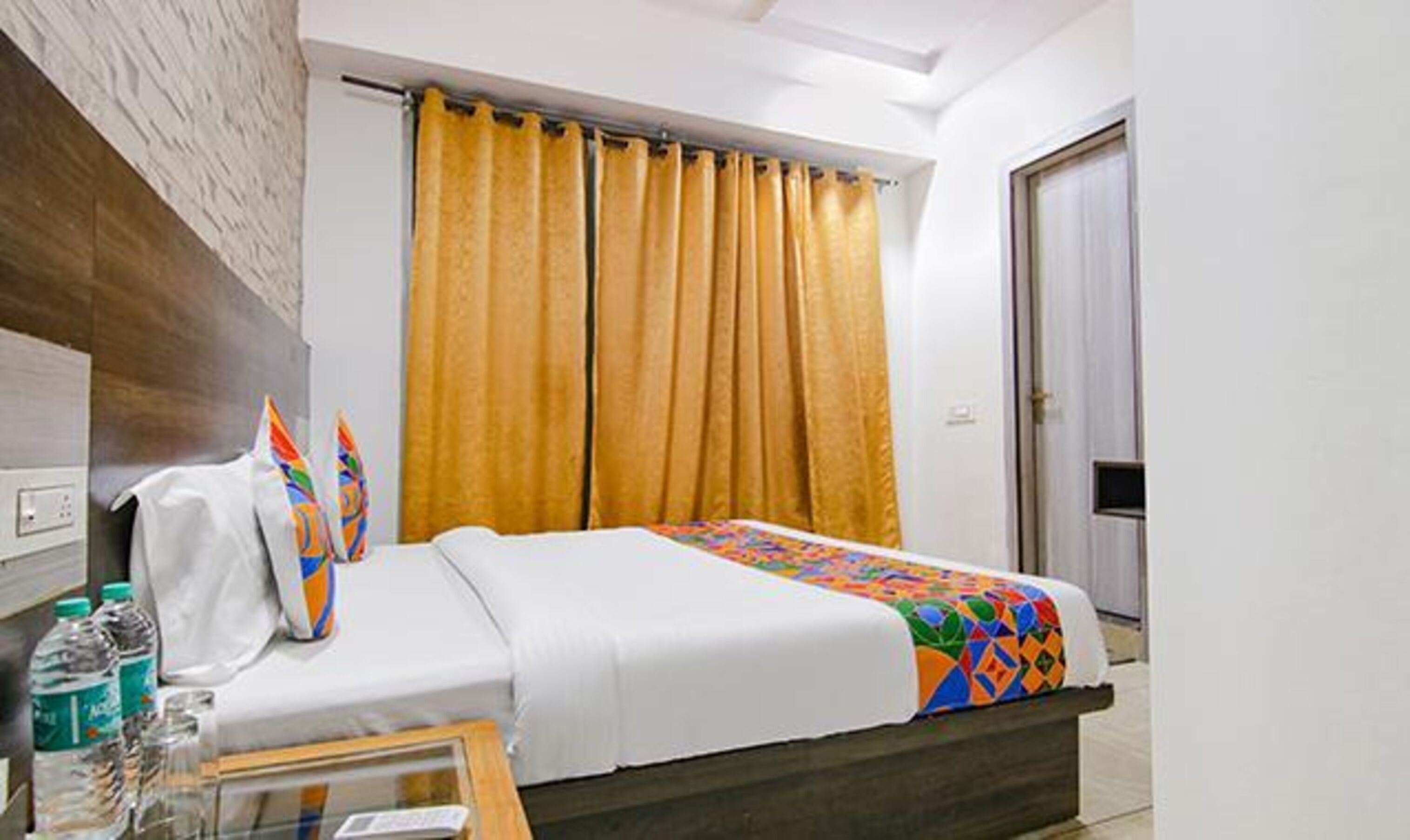 A hotel room with a bed, a wooden headboard, a window with yellow curtains, and a door.