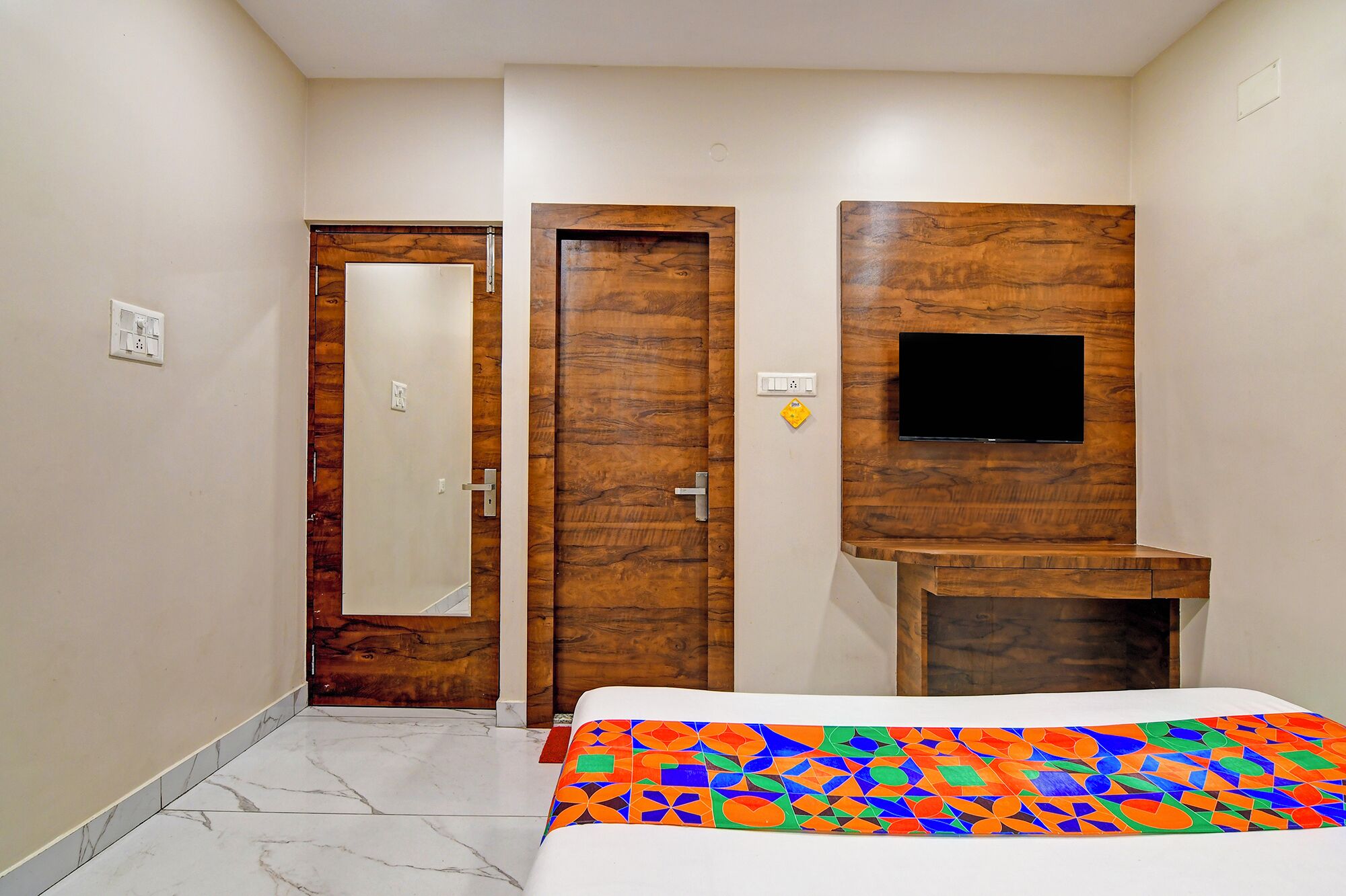 A hotel room with a bed, a TV mounted on a wooden panel, a door with a mirror, and a small desk.