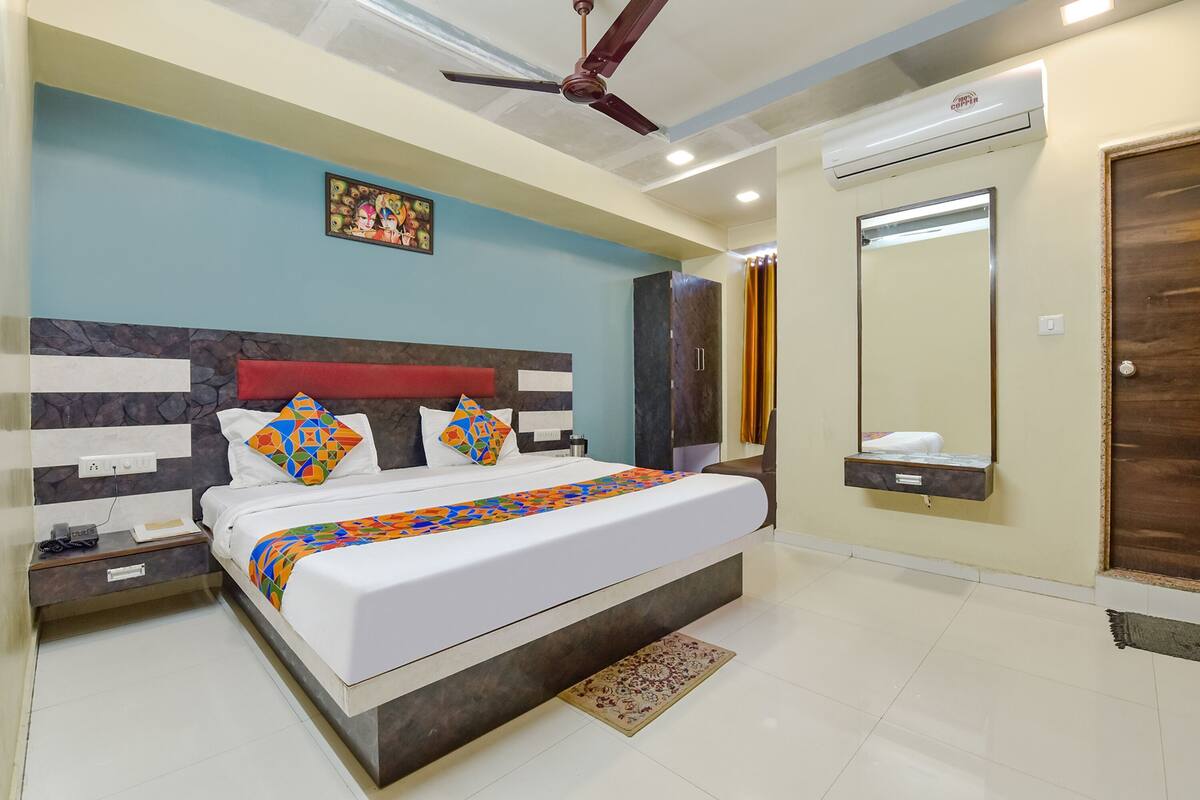 A bedroom with a bed, nightstand, bedside table, wall-mounted air conditioning unit, ceiling fan, and a mirror.