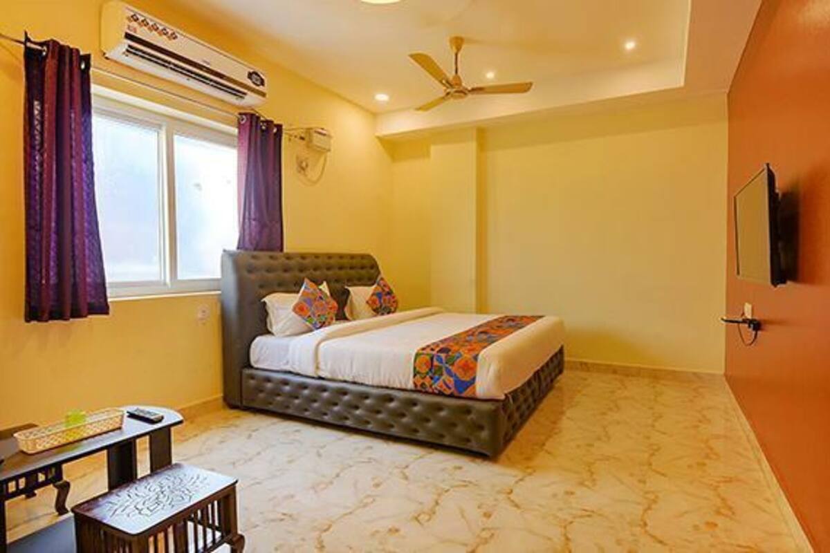 A bedroom with a bed, an air conditioner, a ceiling fan, a window, and a TV mounted on the wall.