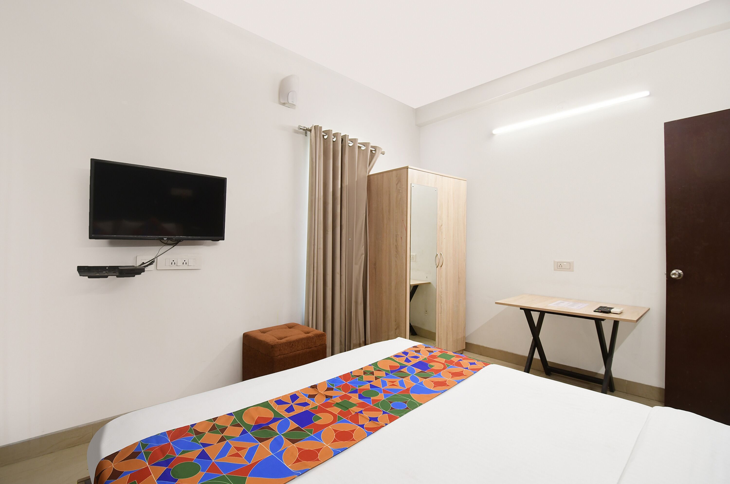 A hotel room with a bed, a TV mounted on the wall, a wooden wardrobe, a small table, and a curtain.