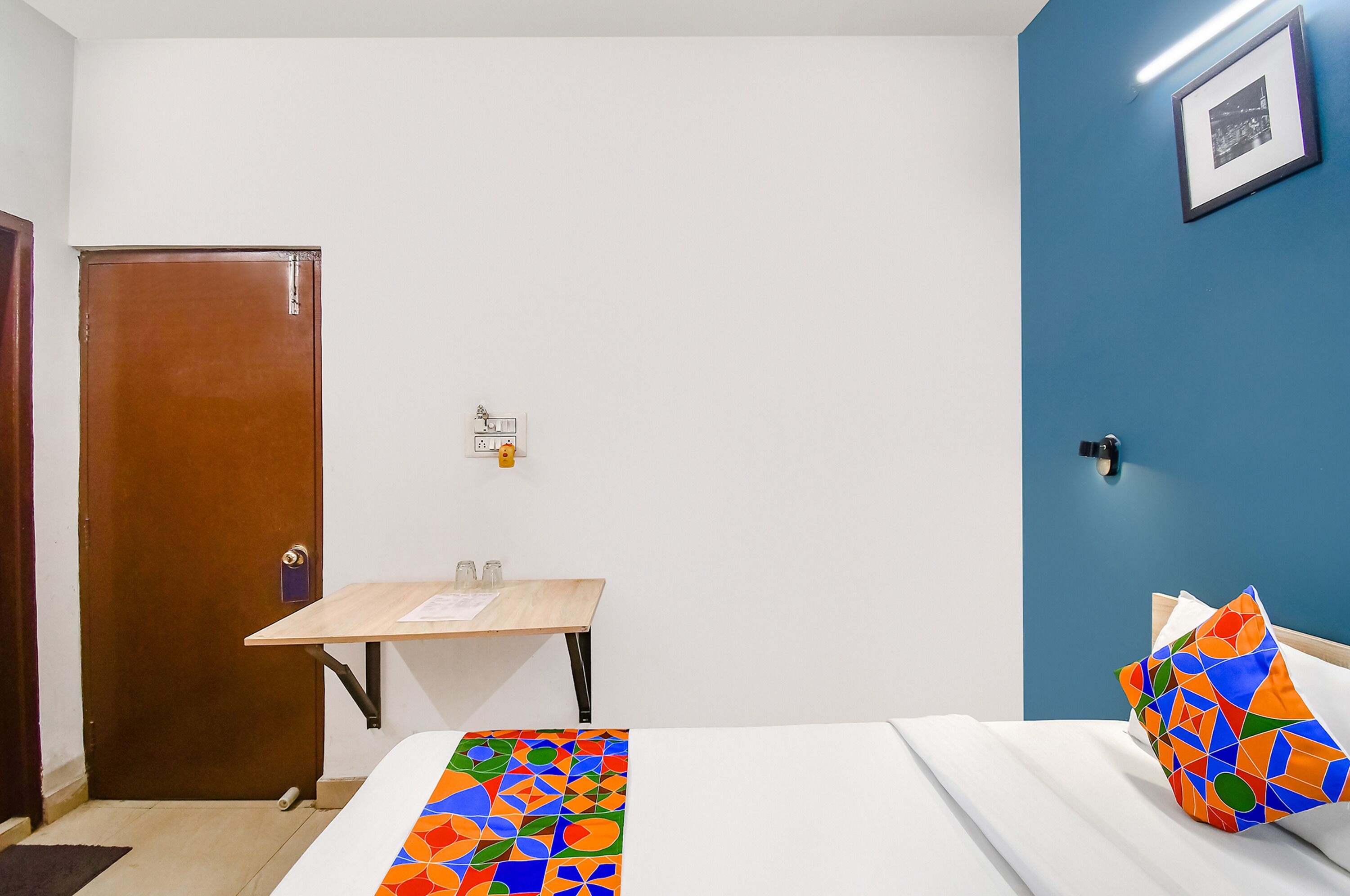 Habitación de hotel con puerta de madera, una mesita con vasos, cama con almohadas de colores y un cuadro en la pared azul.