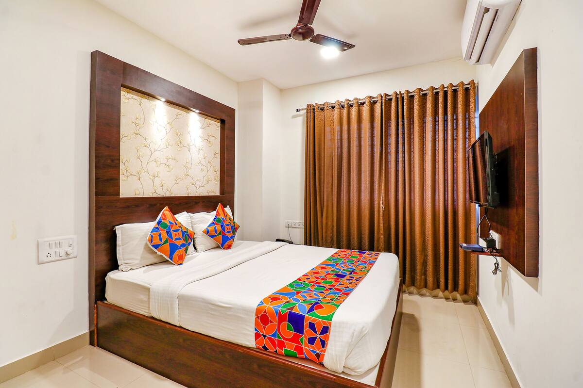A hotel room with a bed, a ceiling fan, an air conditioner, a TV mounted on the wall, and a curtain.