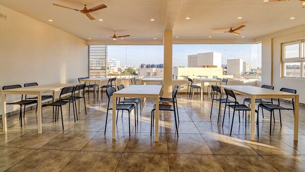 A spacious indoor area with multiple tables and chairs, ceiling fans, and large windows offering a city view. - FabHotel Rotano Grand Marathahalli (Bengaluru)