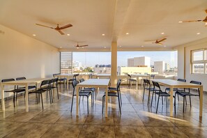 A spacious indoor area with multiple tables and chairs, ceiling fans, and large windows offering a city view.