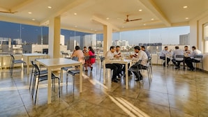 A spacious dining area with high ceilings and large windows offering city views. Tables are arranged for groups, and the floor is tiled. - FabHotel Rotano Grand Marathahalli (Bengaluru)