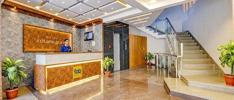 A hotel reception area with a modern design, a reception desk, a TV screen, and a staircase leading to upper floors.