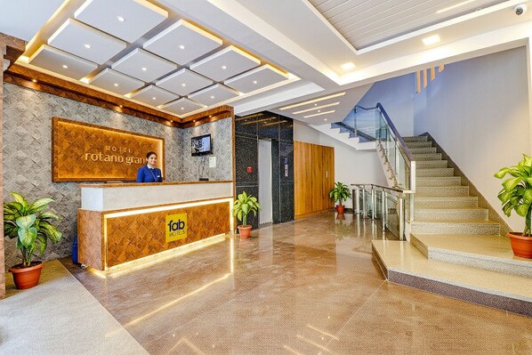 A hotel reception area with a modern design, a reception desk, a TV screen, and a staircase leading to upper floors.
