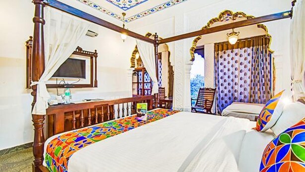 Una cama con dosel y un edredón colorido, un televisor sobre un atril de madera y una ventana con cortinas.