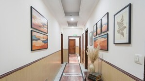 A hallway with framed artwork on the walls, a carpeted floor, and a wooden bench with books and a vase. - Via De Gold I (New Delhi)